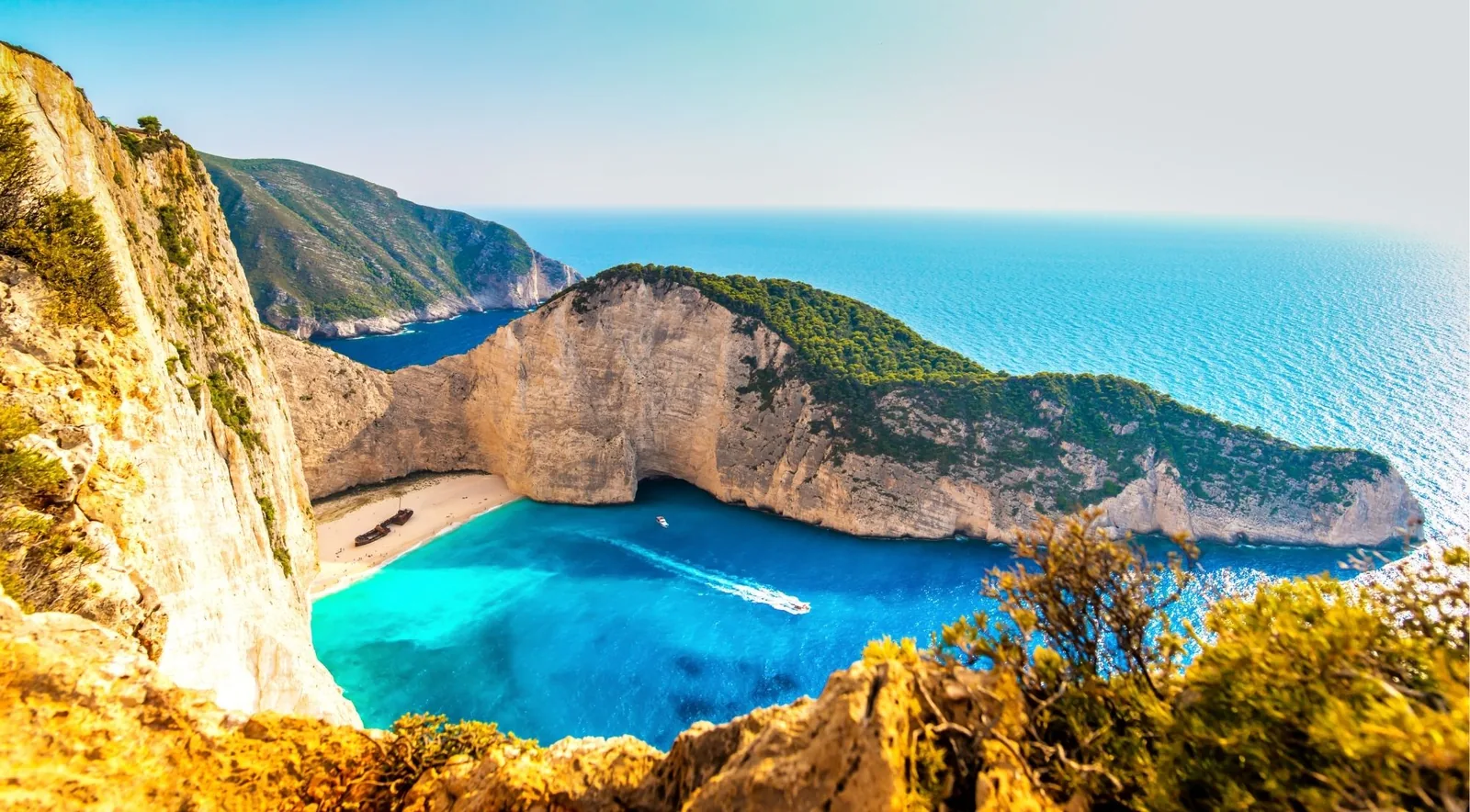 Navagio bay seen from the clifftop viewpoint, the wreck a small dark shape on the sand far below.