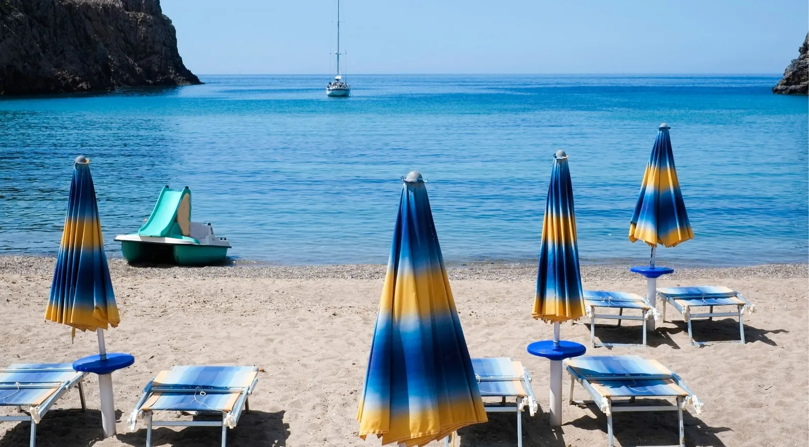 A sheltered sandy cove between rocky headlands, furled blue and yellow umbrellas and loungers on the sand, a pedalo at the water's edge and a yacht anchored offshore
