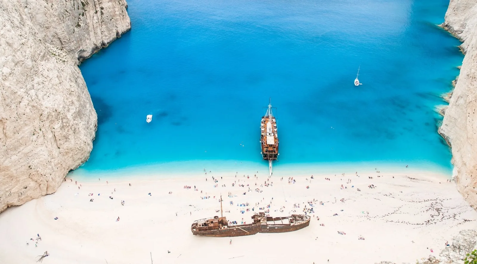 Navagio bay from the clifftop: the rusted wreck on white sand, the bay full of swimmers and a large tourist boat anchored close to the shore