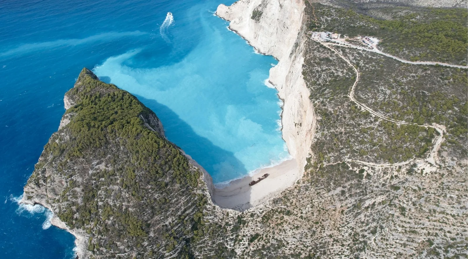 Aerial view of Navagio bay showing the clifftop car park and the pale track leading to it.
